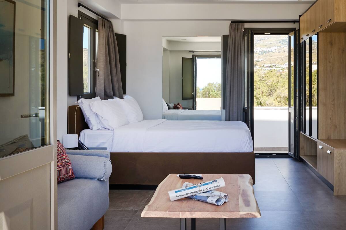 Un hotel moderno con una cama grande, un sofá, una pequeña mesa con periódicos y vistas al exterior.