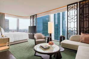 Una habitación de hotel moderna con una cama grande, un sofá, una mesita redonda con libros y flores, un televisor y vistas panorámicas de la ciudad a través de amplias ventanas.