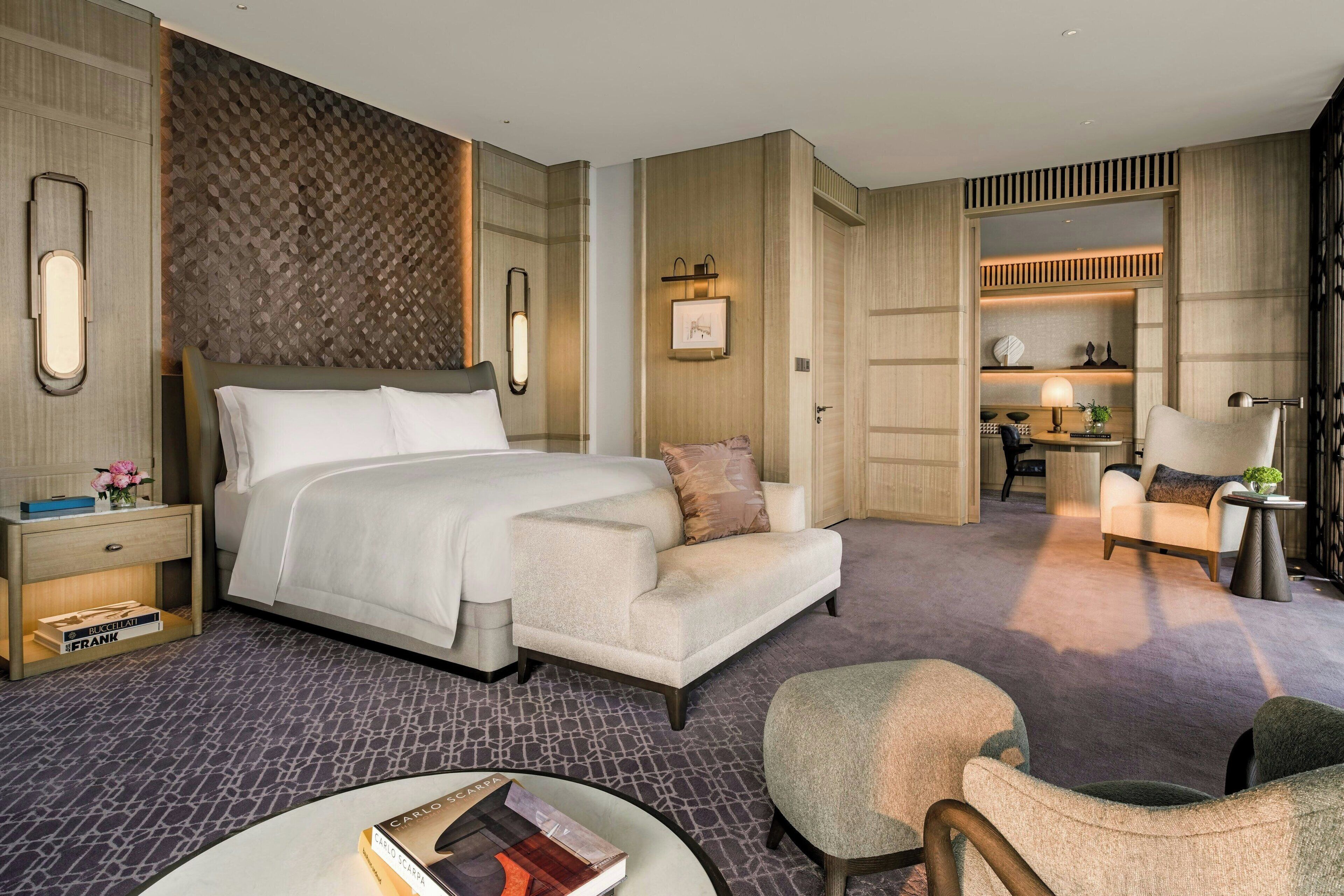 Una habitación de hotel moderna con una cama grande, un sofá, una mesita y una silla. Se ve un buró con libros y una lámpara. La habitación tiene paneles de madera y un papel tapiz estampado.
