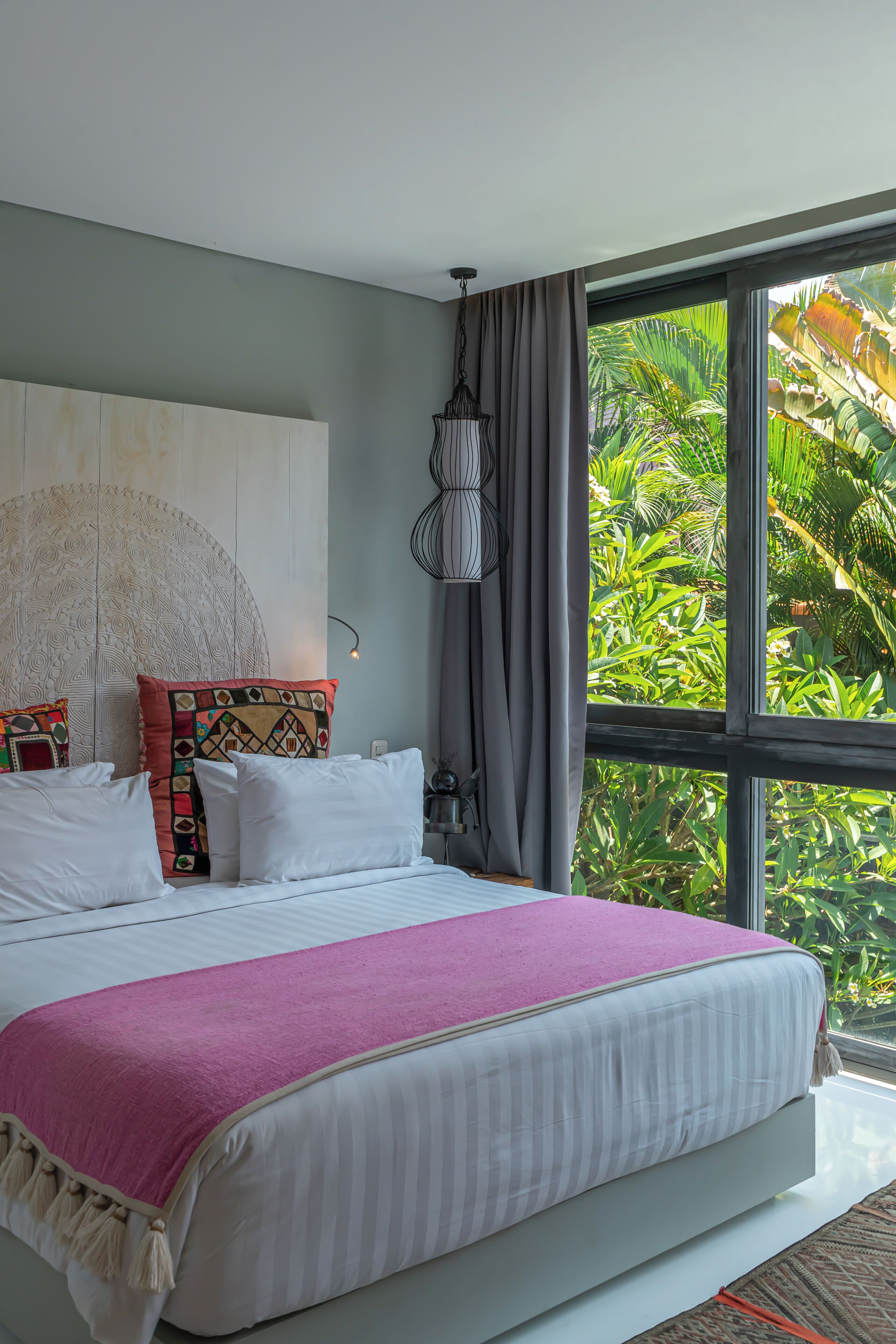A bedroom with a large bed, a decorative headboard, and a view of lush greenery through a large window.