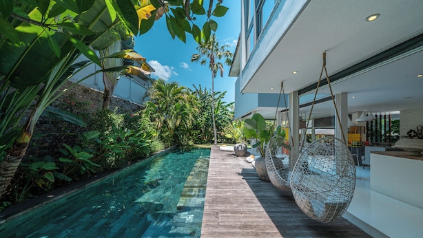 Un área de piscina moderna al aire libre con cubierta de madera, rodeada de exuberante follaje y un cielo azul despejado.
