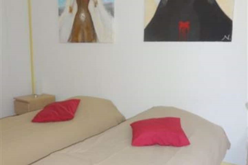 Ein Zimmer mit zwei Betten, jedes mit einem roten Kissen, einem kleinen Nachttisch aus Holz und einem gerahmten Bild an der Wand.