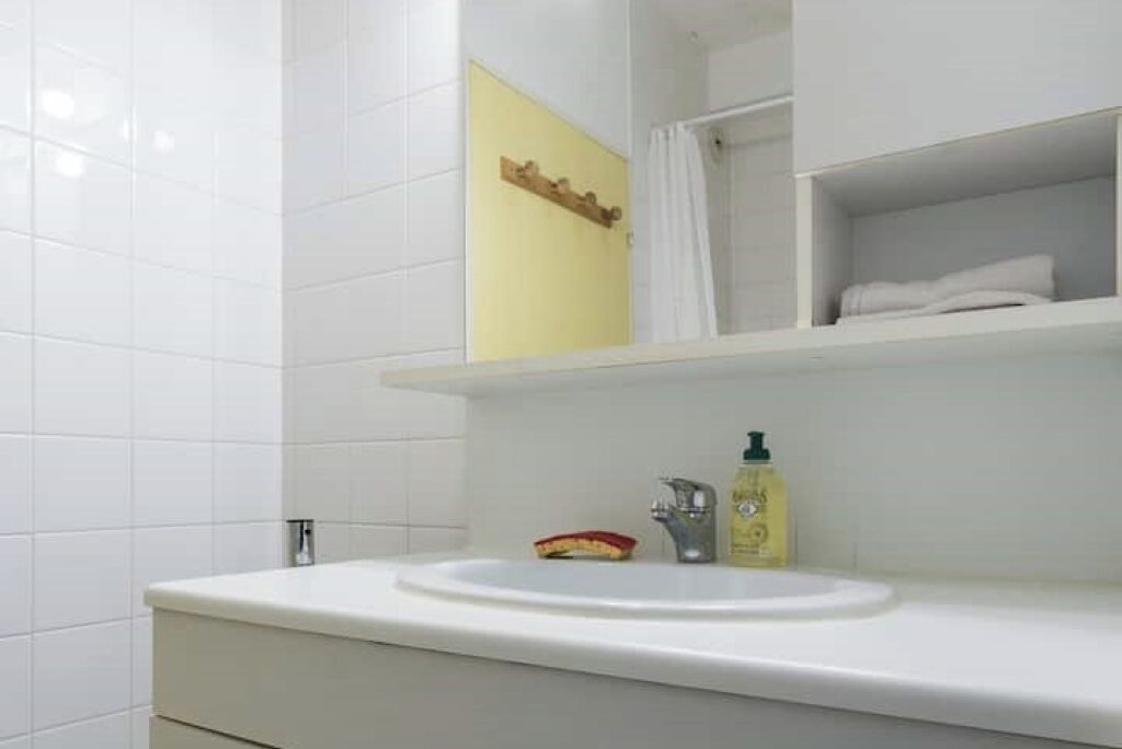 A bathroom with white tiles, a white sink, a yellow wall, and a shelf with a towel.