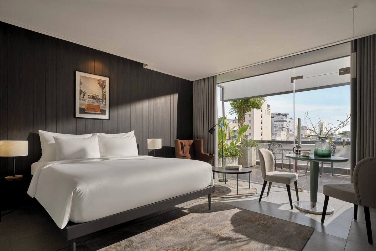 A modern hotel room with a large bed, a small table with chairs, a balcony with a view of the city, and a framed picture on the wall.