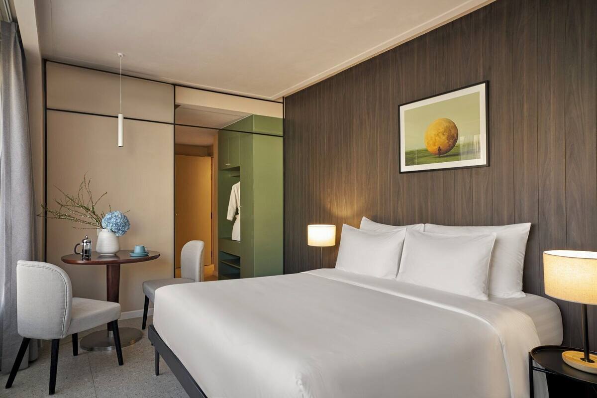 A hotel room with a large bed, bedside lamps, a desk with a chair, a small table with a vase and cup, and a green wardrobe.