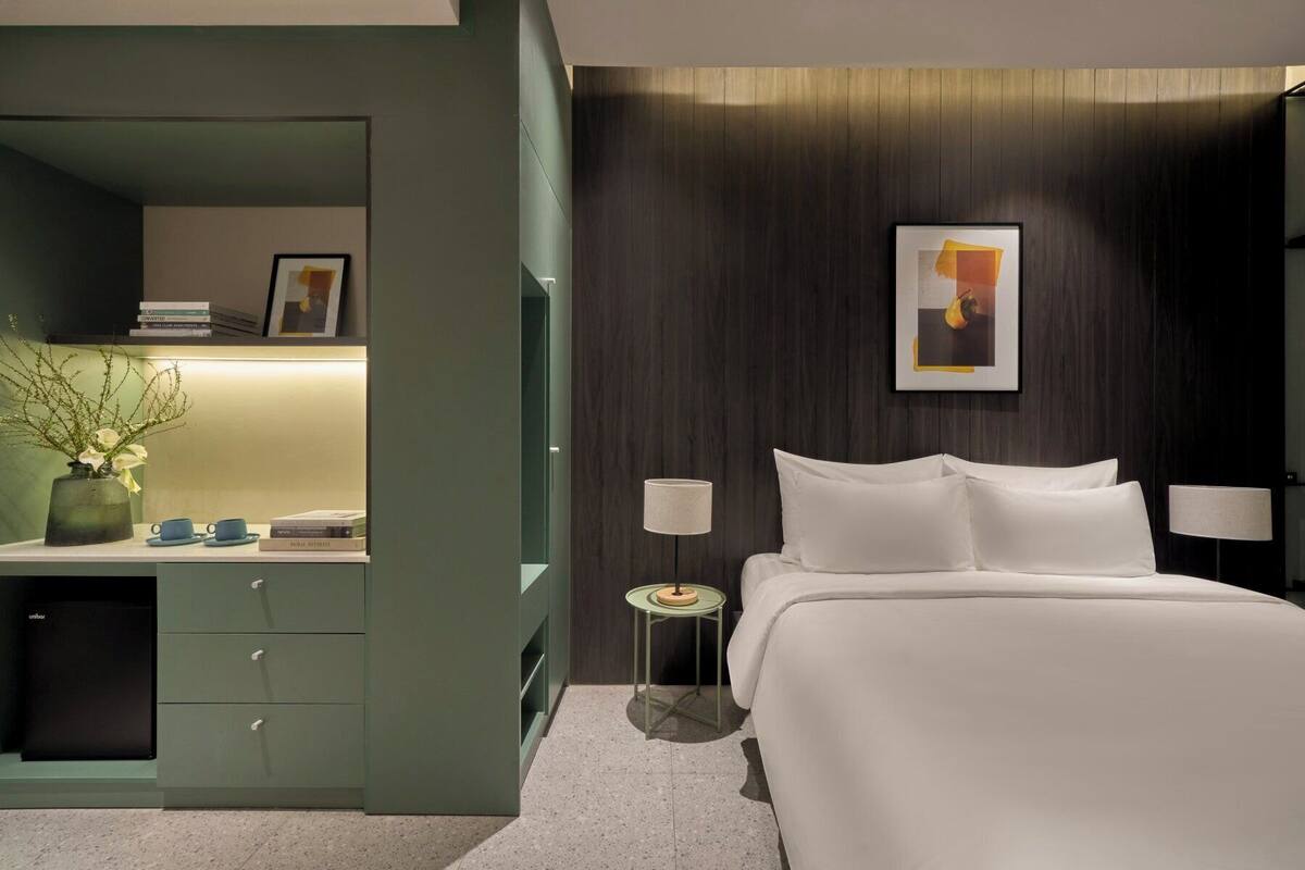 A modern hotel room with a bed, bedside lamps, a small table, a desk with a lamp, and a picture on the wall.