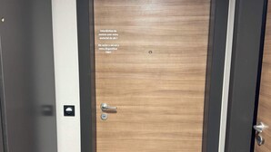 A wooden door with a metal handle and a keyhole, a small mat on the floor, and a wall-mounted light switch. - Magnificent new 60m2 apartment in an exceptional location! (Samoëns)