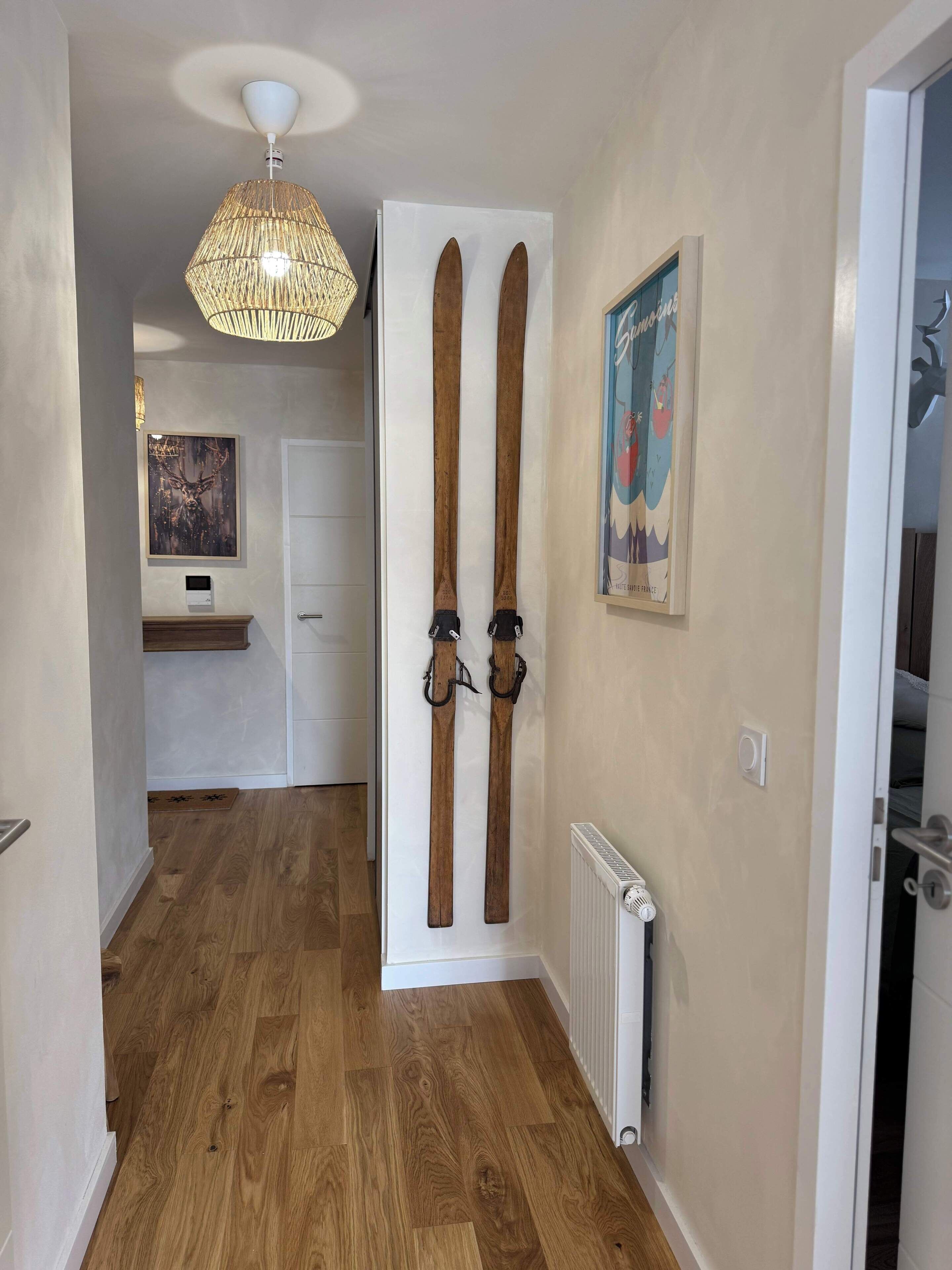 A hallway with wooden flooring, a white door with ski bindings, a poster, and a ceiling light fixture.
