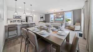 Une cuisine moderne équipée d’appareils électroménagers en acier inoxydable, un coin repas avec une longue table pouvant accueillir six personnes, et un salon avec un canapé et un téléviseur.