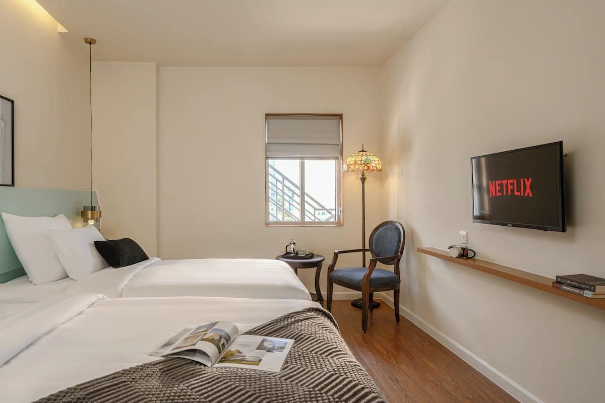 A hotel room with two beds, a TV displaying 'NETFLIX', a chair, a small table, and a window with a view of buildings.