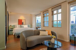 A modern hotel room with a large bed, a grey sofa, a small table, and large windows offering city views. - M Village Hotel Tao Đàn Park, a brand of Modern Village Lifestyle (Ho Chi Minh City)