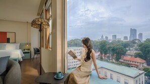 Habitación de hotel con un ventanal amplio que ofrece vistas a la ciudad, una cama, un sillón para leer y una mesa con una taza.