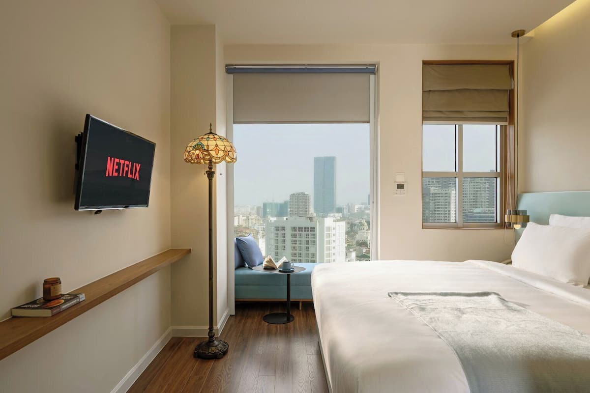 A hotel room with a bed, a TV mounted on the wall, a floor lamp, a window with a view of the city, and a bedside table with a book and a cup.