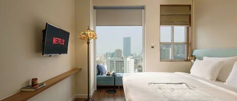 A hotel room with a bed, a TV mounted on the wall, a floor lamp, a window with a view of the city, and a bedside table with a book and a cup.