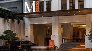 Una entrada de hotel moderna con un gran letrero con la letra 'M', una escalera que conduce al vestíbulo y un SUV negro estacionado afuera.