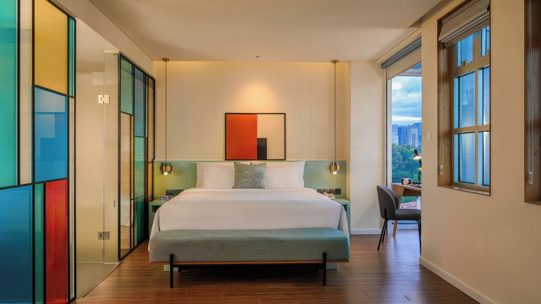 A modern hotel room with a large bed, a desk, and a view of the city through large windows.