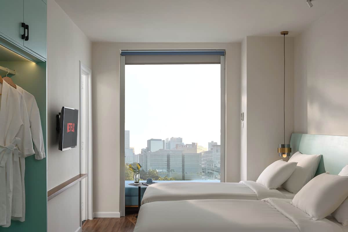 A modern hotel room with a large bed, a wardrobe, a desk with a chair, and a city view through the window.