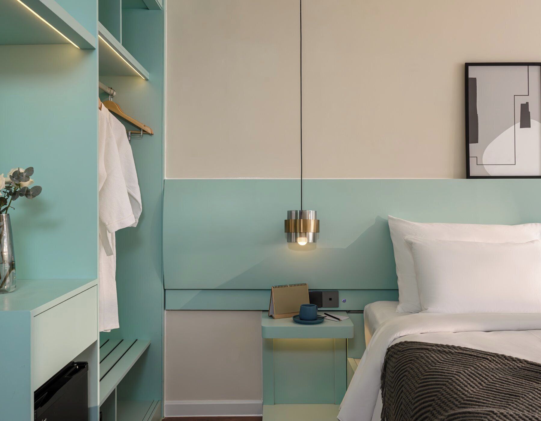 Hotellihuone, jossa on sänky, yöpöytä, työpöytä, vaatekaappi ja seinään kiinnitetty valaisin.