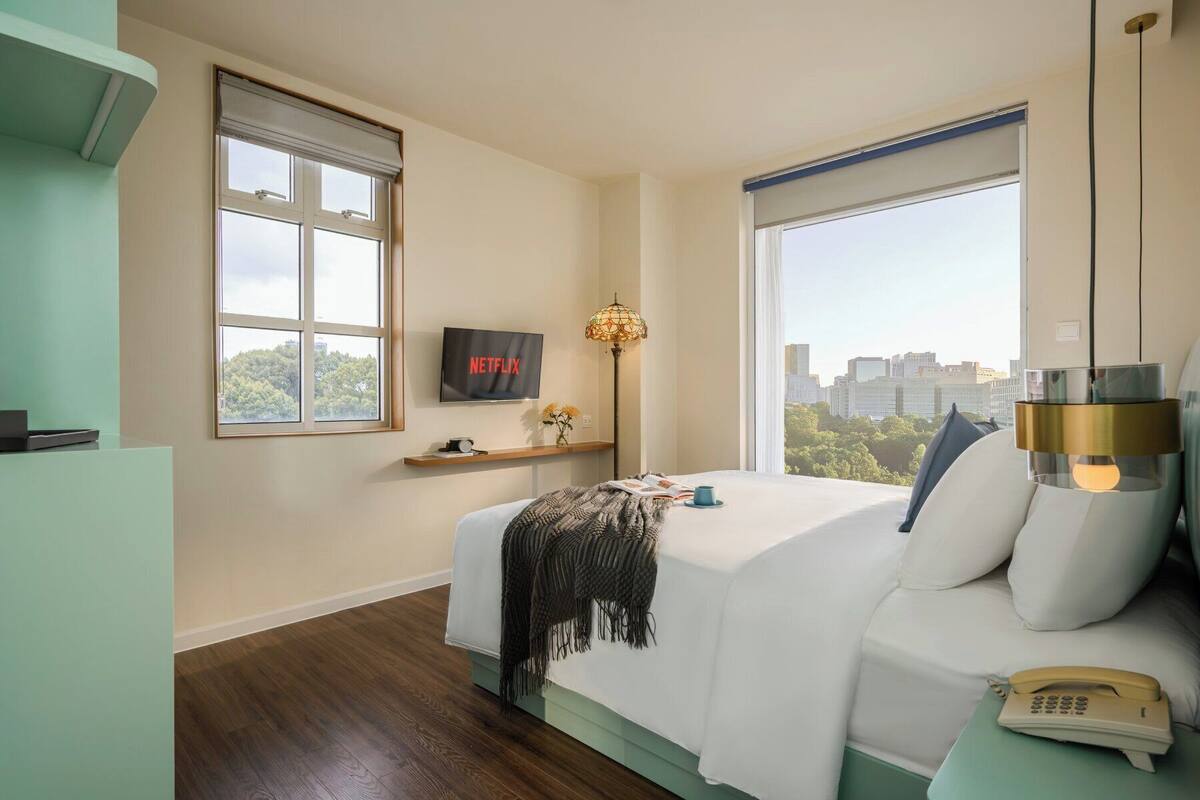 A modern hotel room with a bed, a TV, a lamp, and a city view.
