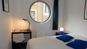 Een hotelkamer met een rond raam, een bureau met stoel, een bed met blauwe kussens en een nachtkastje met een lamp.
