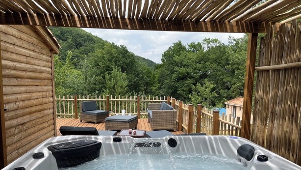 A hot tub with a wooden deck and wicker furniture, surrounded by a wooden fence and lush greenery. - Chalet/Spa and private lake (CHALABRE)