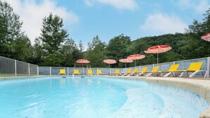 A swimming pool with yellow lounge chairs and red umbrellas, surrounded by a fence and trees. - Chalet/Spa and private lake (CHALABRE)