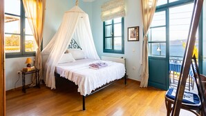 A bedroom with a bed, a bedside table with books, a window with curtains, and a view of the sea.