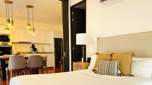 A hotel room with a large bed, a dining area with chairs, a kitchen, and a bedside lamp.