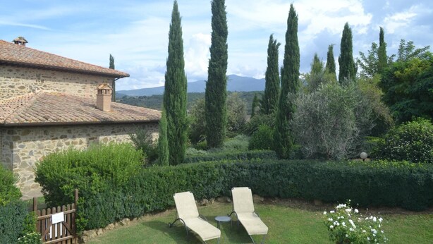 A stone building with a tiled roof, two lounge chairs, and a well-maintained garden with tall trees and hedges.