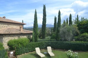 A stone building with a tiled roof, two lounge chairs, and a well-maintained garden with tall trees and hedges.