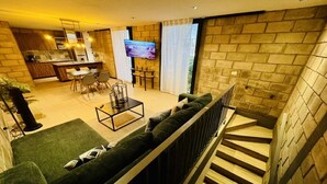 A modern living room with a green sofa, a coffee table, and a TV. There is a kitchen in the background and a staircase with wooden steps.