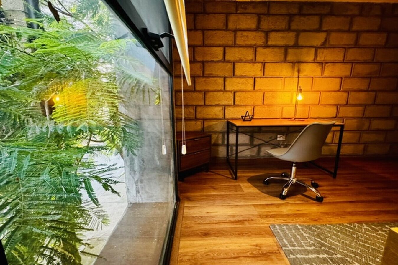 A room with a brick wall, a wooden floor, a desk with a chair, and a large window with a plant.