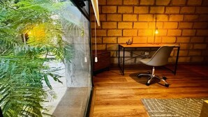 A room with a brick wall, a wooden floor, a desk with a chair, and a large window with a plant.