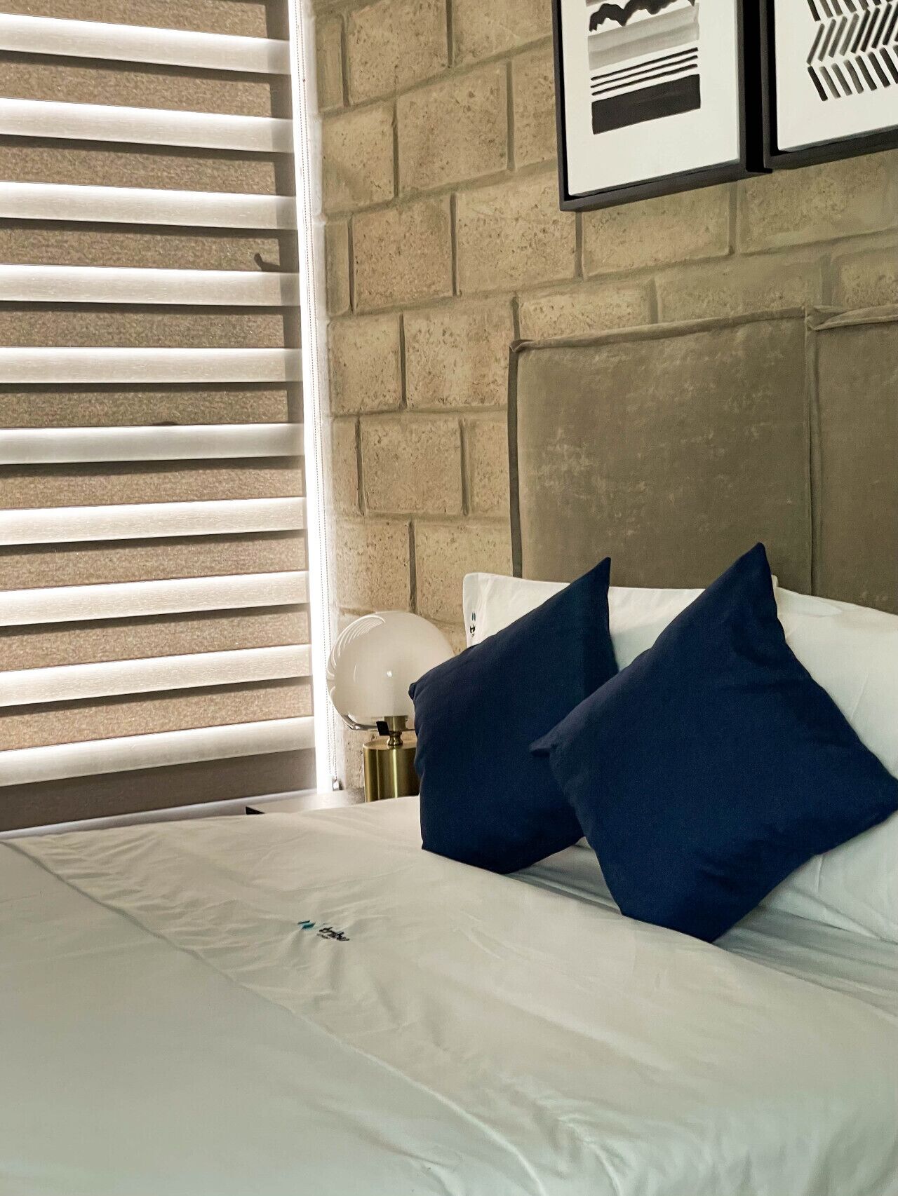 Ein Bett mit weißer Bettwäsche und zwei dunkelblauen Kissen, eine Nachttischlampe und eine Backsteinwand mit gerahmten Kunstwerken.