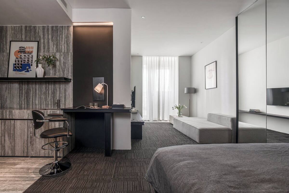 A modern hotel room with a dark wood accent wall, a desk with a chair, a sofa, a bed, and a large window with sheer curtains.