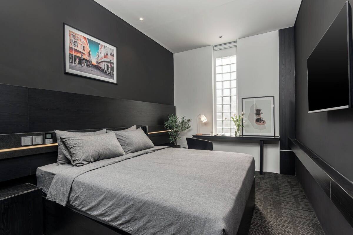 A modern hotel room with a large bed, a desk with a lamp, a window with blinds, and a framed picture on the wall.