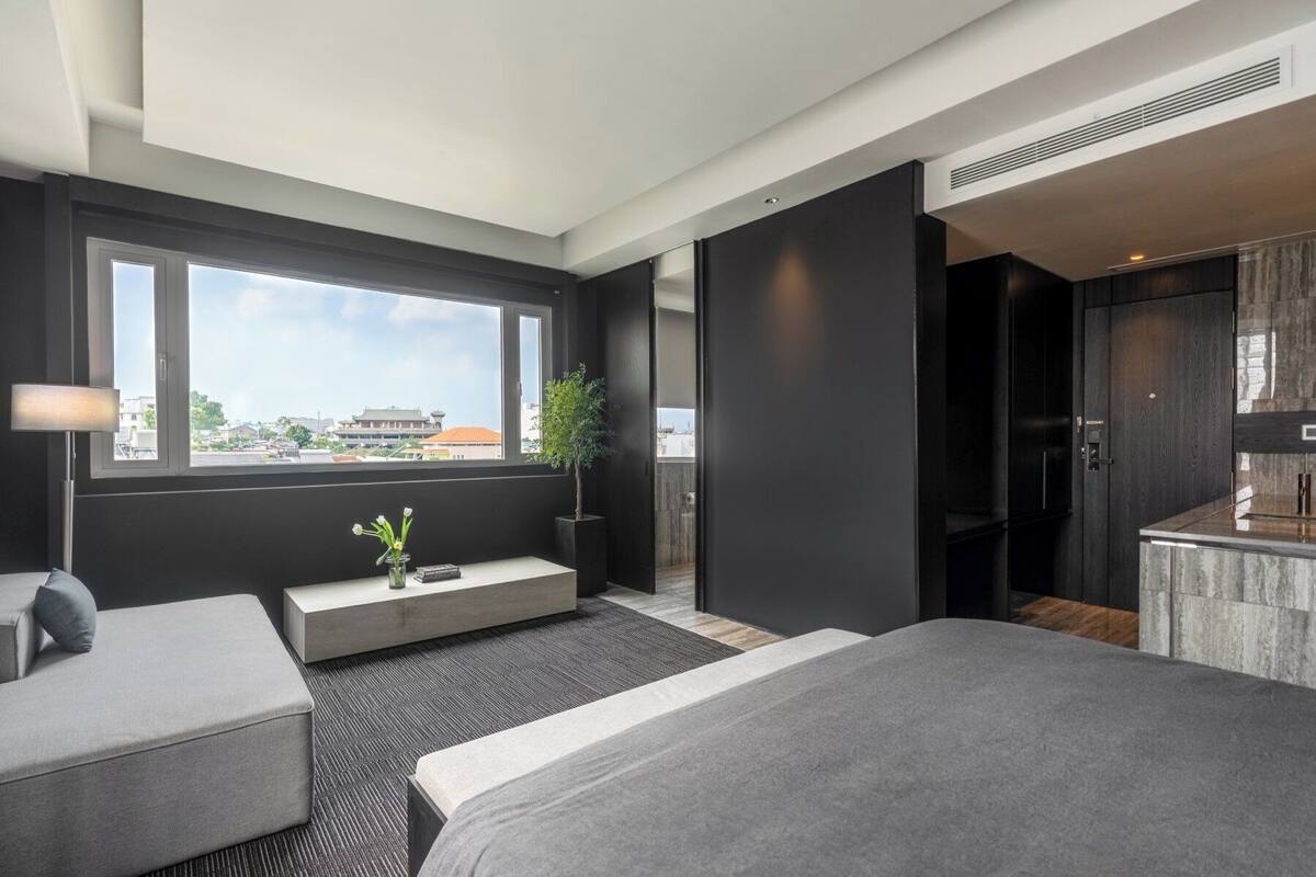 A modern hotel room with a large bed, a seating area, a small table with a vase, a potted plant, and a view of the city through a large window.