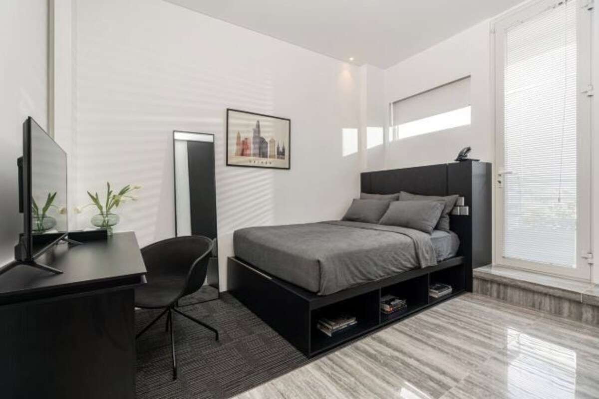 A modern bedroom with a bed, a desk, a chair, a TV, a mirror, and a window.