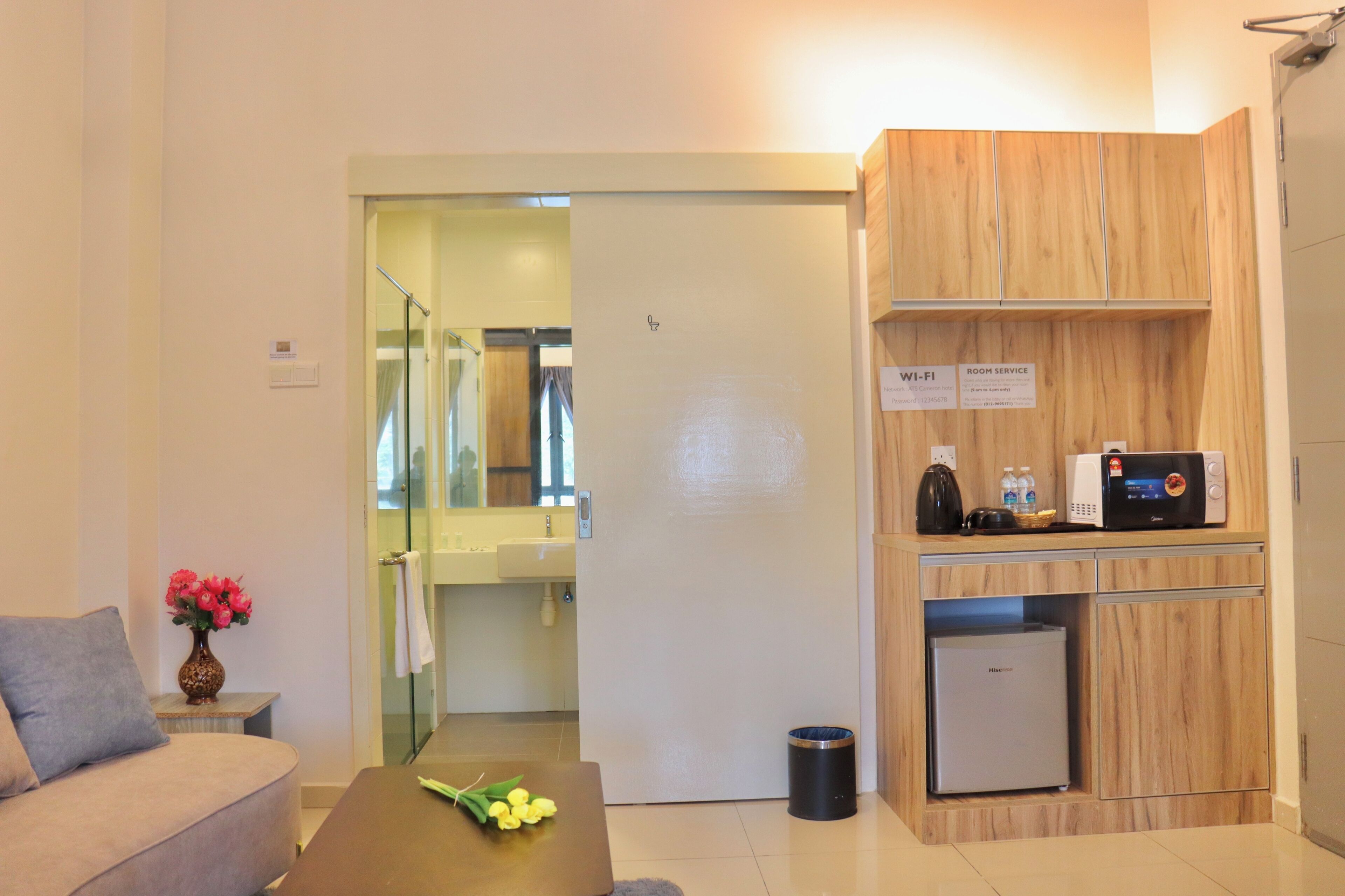 Habitación de hotel moderna con sofá, una mesita con flores, kitchenette con microondas y un cartel que indica servicio de Wi-Fi y habitaciones.