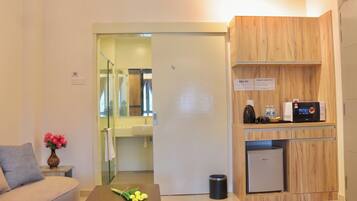 Habitación de hotel moderna con sofá, una mesita con flores, kitchenette con microondas y un cartel que indica servicio de Wi-Fi y habitaciones.