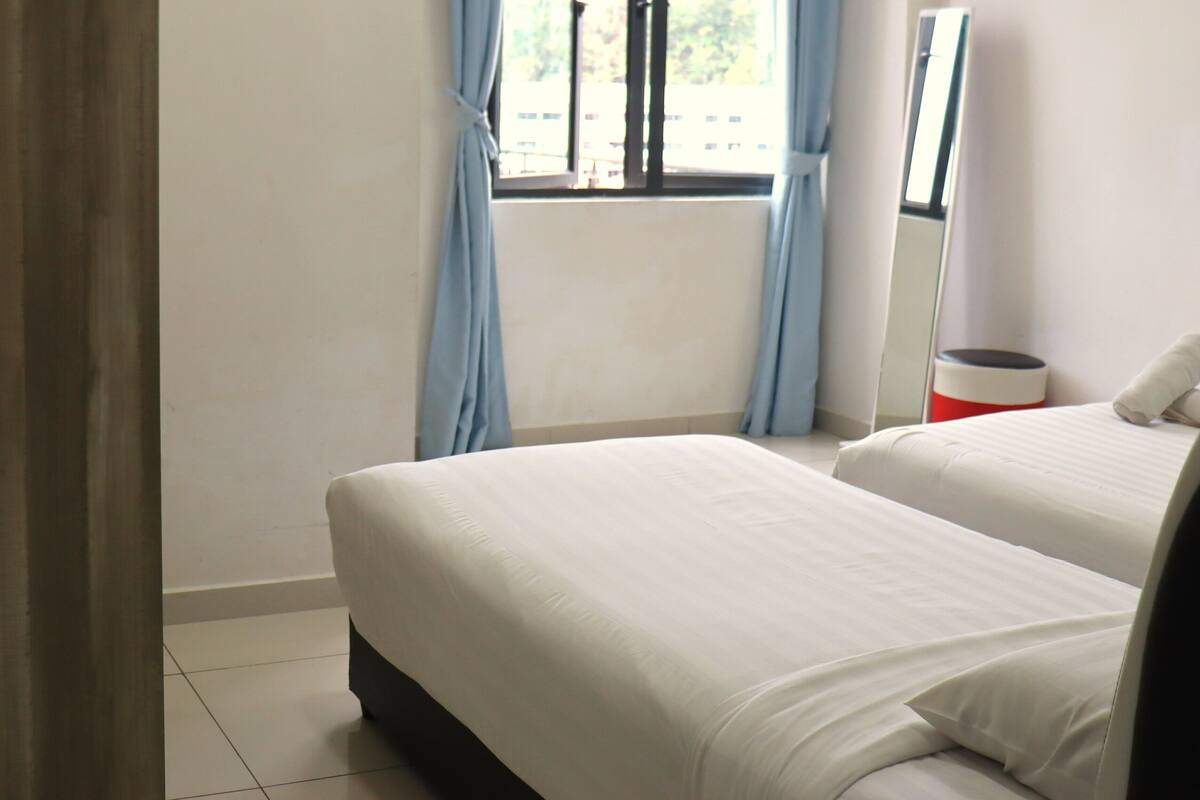 A hotel room with two beds, a window with curtains, a mirror, and a trash bin.