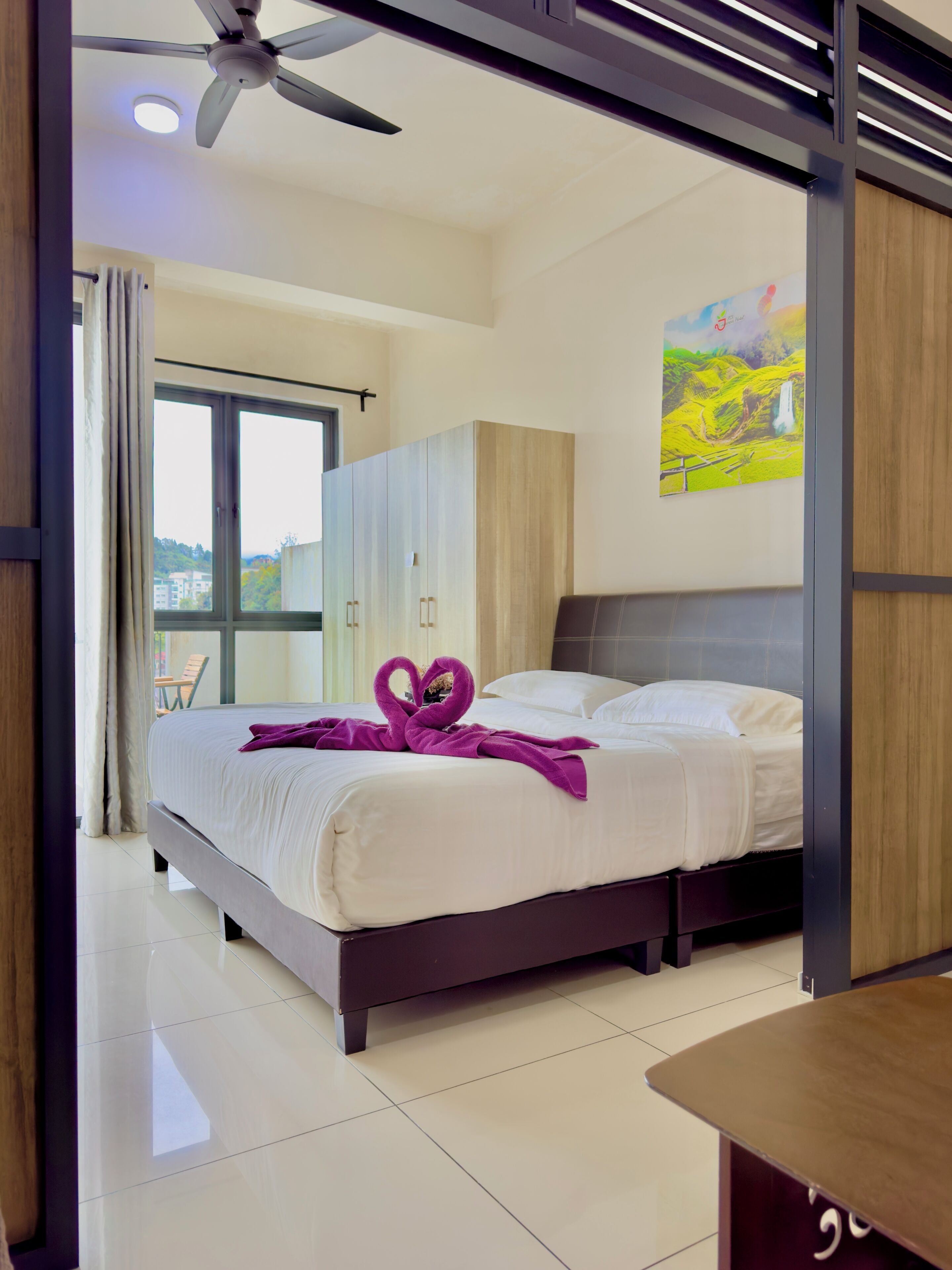A hotel room with a bed, a ceiling fan, a window with curtains, and a painting on the wall.