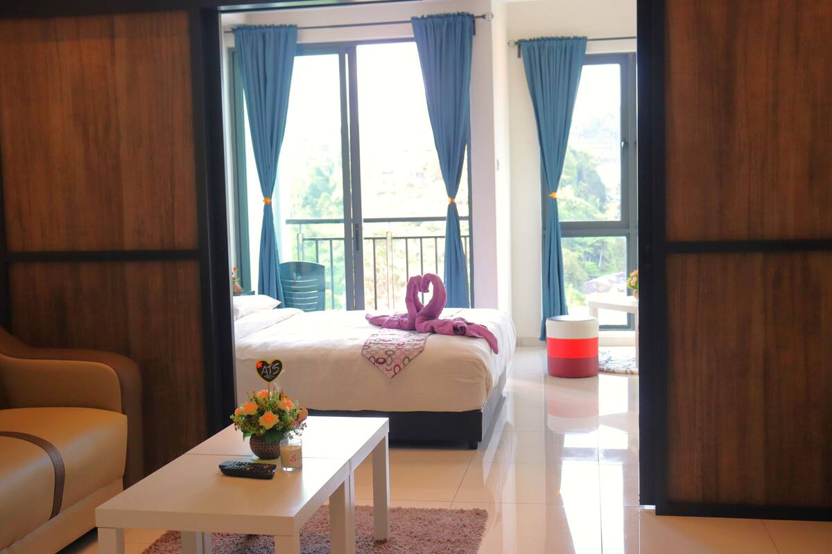 A hotel room with a bed, a sofa, a table with a vase of flowers, and a view of the outside through large windows.