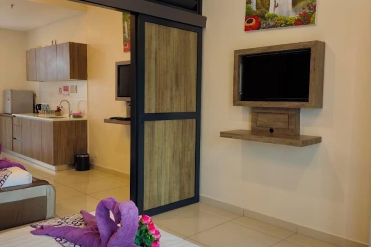 A modern hotel room with a bed, a TV mounted on the wall, a kitchen area visible through a sliding door, and a small shelf above the TV.