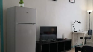 Et lille værelse med et hvidt køleskab, et fladskærms-tv på et sort stativ, et skrivebord med lampe og en stol.