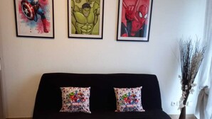 Ein Wohnzimmer mit einer schwarzen Couch, zwei Kissen mit Marvel-Helden-Motiven und einem Teppich im Marvel-Design.