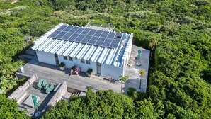 A modern house with a solar panel roof, surrounded by lush greenery and a beach view.