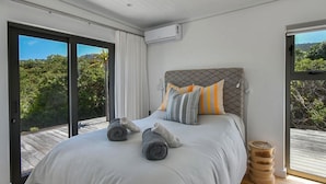 A modern bedroom with a large bed, a view of greenery through the window, and a wooden floor.