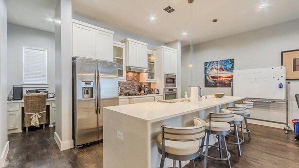 A modern kitchen with white cabinetry, a central island with bar stools, a stainless steel refrigerator, and a built-in oven. - Modern 2 BR, 4 Story Smarthome with Rooftop Hot Tub, Game Room and Skyline Views (Dallas)