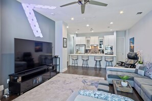 A modern living room with a flat-screen TV, a ceiling fan, and a kitchen area with a bar.